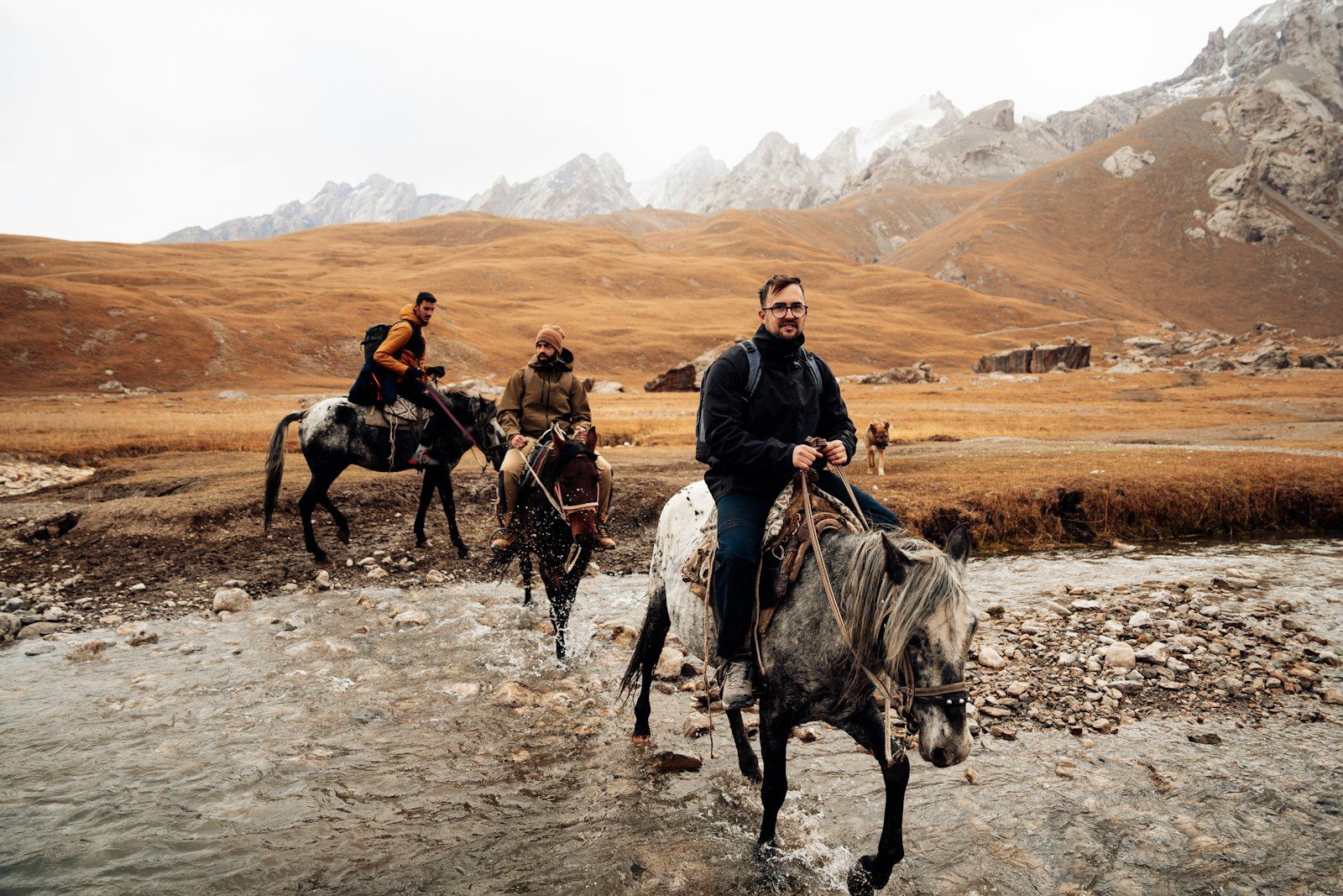 Three riders and a dog crossing a shallow braided river below jagged snow-dusted peaks.