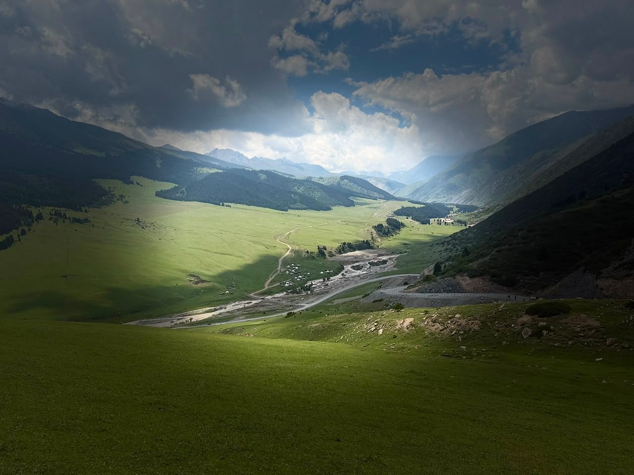 Sunlight breaking through storm clouds over a green valley, riverbed, road, and small settlement.