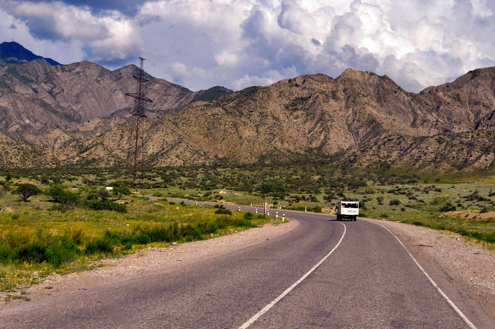 A minibus travels a paved road below dry, ridged mountains under a partly cloudy sky.