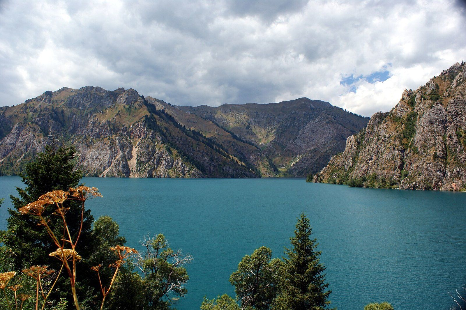 The turquoise water of Sary-Chelek lake ringed by steep forested mountains under storm clouds.