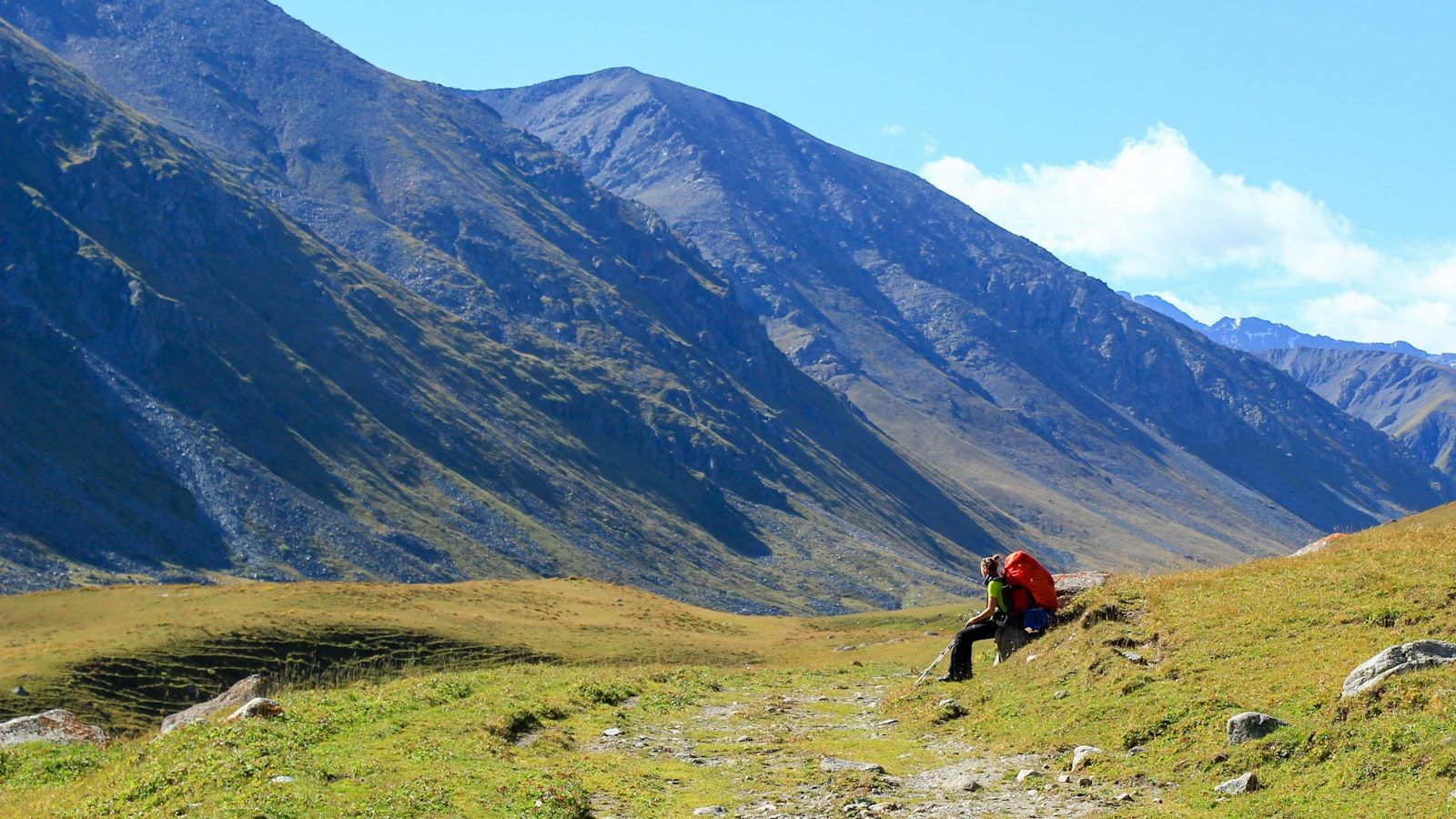 A trekker resting on a mountain trail with a loaded backpack and rolled sleeping mat, a broad scree-and-grass ridge rising behind.