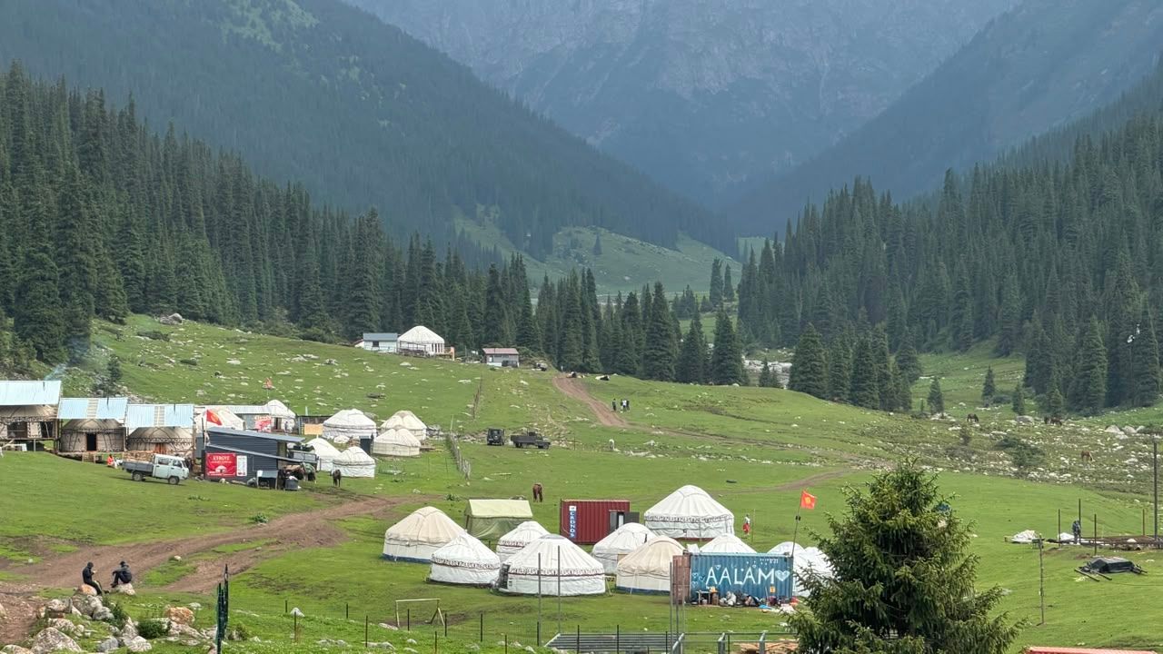 Yurt camp in a forested mountain valley, with vehicles, containers, and a Kyrgyz flag.