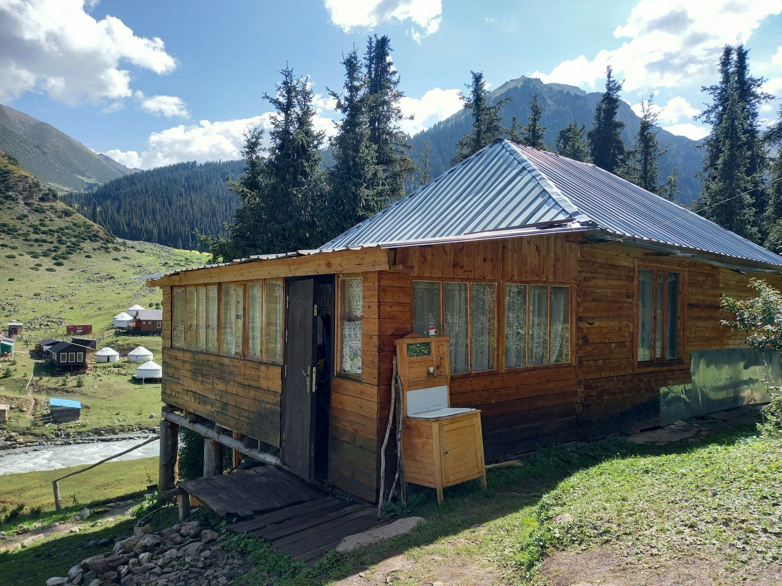 A wooden guesthouse cabin on stilts above a mountain stream, with yurts in the valley below.
