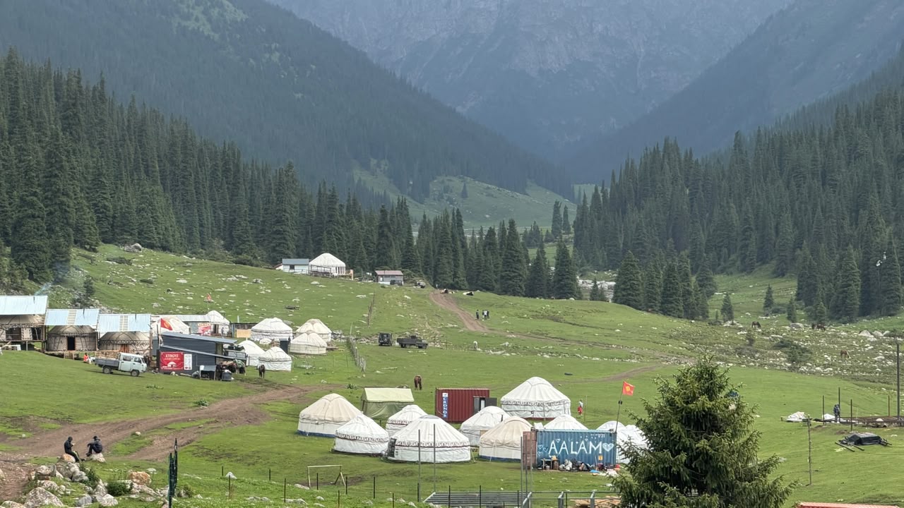 Yurt camp in a forested mountain valley, with vehicles, containers, and a Kyrgyz flag.